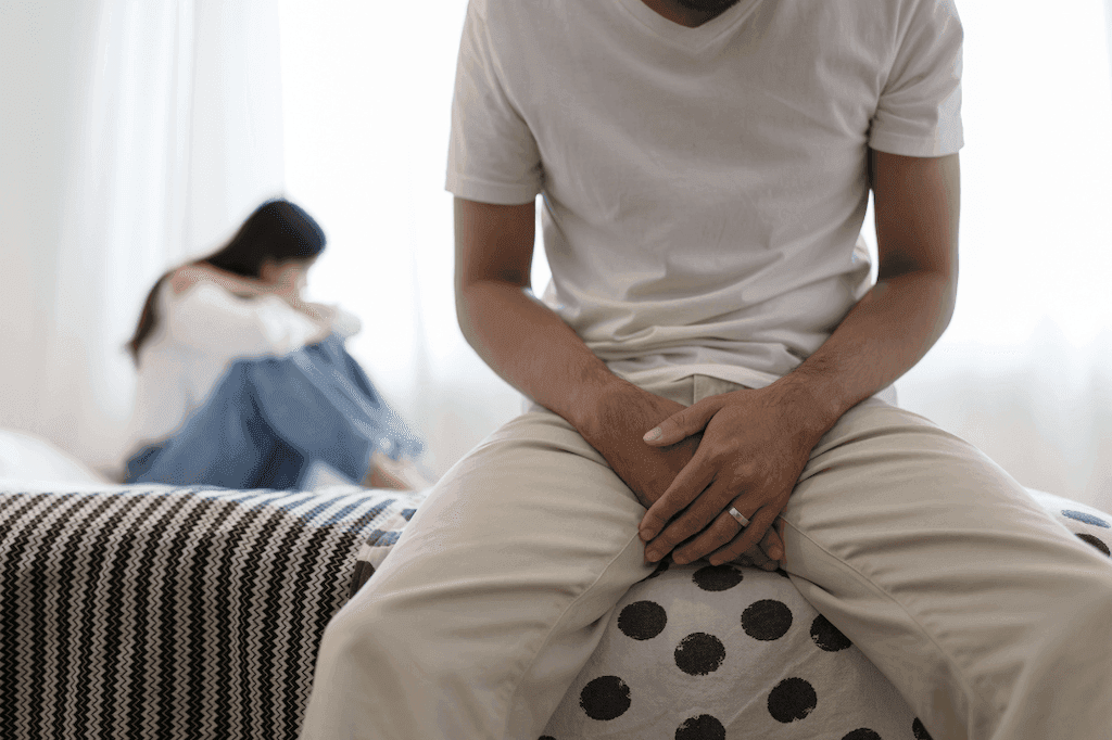 Man sitting on the edge of the bed, in a white shirt and pants, hands covering his crotch. Woman in the background, curled up sitting on the bed, had is down as she embraces herself. 