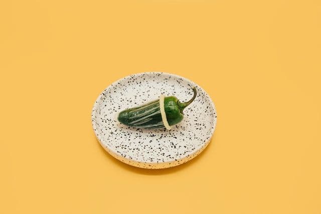 Shishito pepper covered in a condom sitting on top of a black and white speckled dish. Mustard-yellow background. 