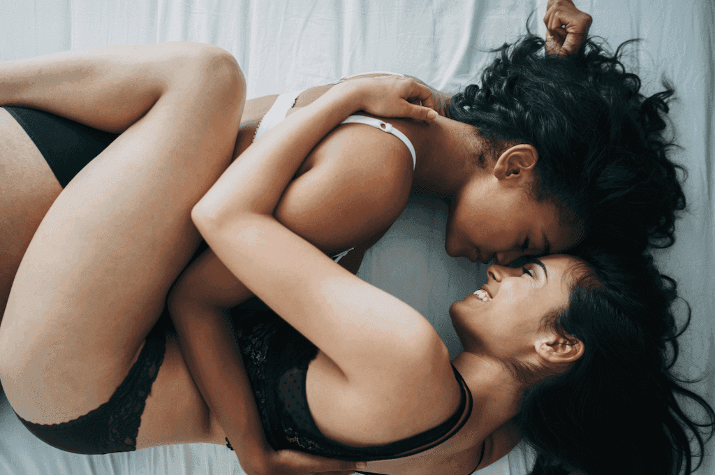 Two women embracing in bed, legs intertwined. Both are in a bra and underwear. They smile at each other, foreheads touching. 